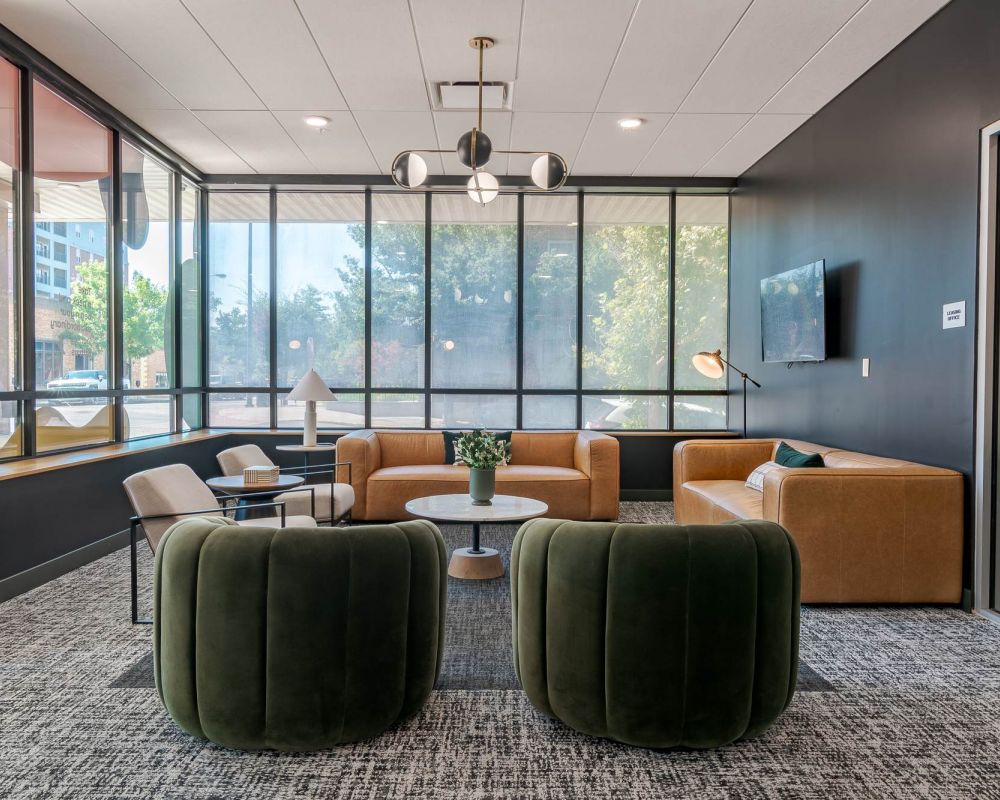 A stylish resident lounge featuring large windows, natural light, and modern seating with green and tan accents, offering a comfortable place to relax or gather at District Flats Columbia.