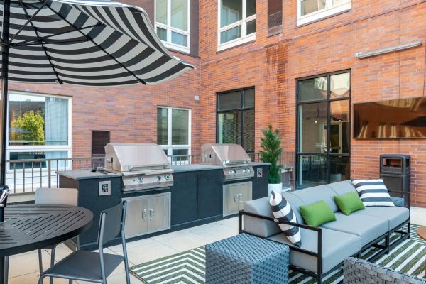 A modern outdoor grill area with shaded picnic tables and stainless steel barbecue stations surrounded by landscaped greenery at District Flats Columbia.